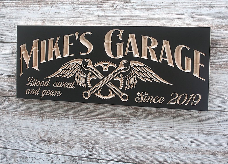 May include: Black wooden sign with the text "Mike's Garage" in a decorative font. The sign also features a pair of crossed wrenches with wings and gears, and the text "Blood, sweat, and gears" and "Since 2019".