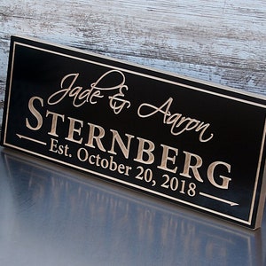 May include: Black wooden sign with a gold inscription that reads "Jade & Aaron Sternberg Est. October 20, 2018".