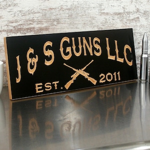 May include: Black wooden sign with the text "J & S GUNS LLC EST. 2011" in a light brown color. Two rifles are crossed below the text. Two bullets are standing upright to the right of the sign.