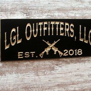 May include: A black wooden sign with gold lettering that reads "LGL Outfitters, LLC Est. 2018". Two gold crossed rifles are centered below the text.