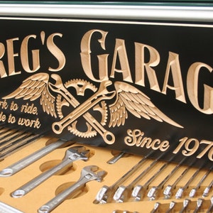 May include: A black wooden sign with the words "Greg's Garage" carved in a light brown wood. The sign also features a pair of crossed wrenches with wings, a gear, and the text "Work to ride, Ride to work" and "Since 1976".