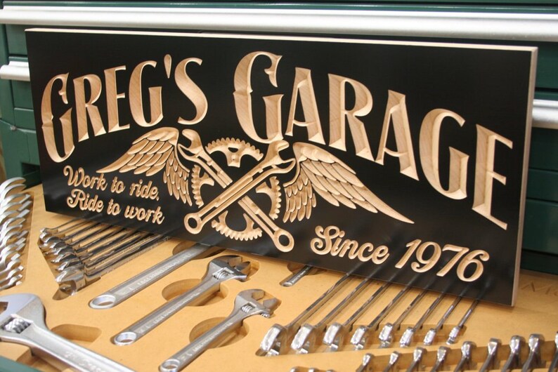 May include: A black wooden sign with the words "Greg's Garage" carved in a light brown wood. The sign also features a pair of crossed wrenches with wings, a gear, and the text "Work to ride, Ride to work" and "Since 1976".
