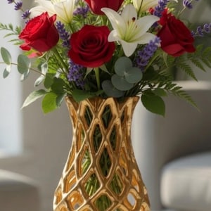 May include: A gold vase with a geometric pattern contains a colourful flower arrangement. The bouquet includes red roses, white lilies, and purple lavender, with green leaves. The vase is on a light surface.
