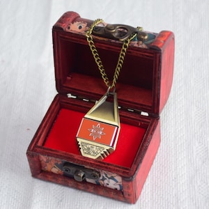 Puede incluir: Un colgante metálico con un centro rectangular naranja y un emblema de sol plateado, suspendido de una cadena dorada dentro de un pequeño cofre de madera rectangular con interior de terciopelo rojo y un cierre metálico frontal.