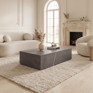 May include: A rectangular, low-profile coffee table made of dark grey stone with subtle white veining. The table features a solid, block-like construction and sits centrally on a textured, cream-coloured area rug in a bright, neutral-toned room.