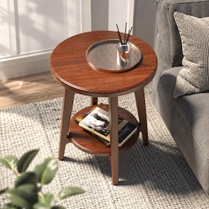 Puede incluir: Una mesa auxiliar redonda de madera con un acabado marrón cálido, que cuenta con una superficie circular y un estante inferior. La parte superior sostiene una bandeja plateada con un difusor de vidrio y varillas negras, y dos libros apilados.