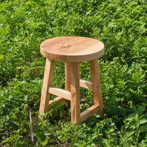 May include: A round wooden stool with a light brown natural grain finish, featuring a circular seat supported by four straight legs connected by horizontal crossbars, showcasing the visible texture and knots of the light-coloured wood.