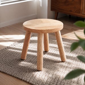 May include: A small, round wooden stool featuring a smooth, light-toned natural wood grain finish. The stool is supported by four sturdy, cylindrical legs that extend outward from the circular seat, providing a stable base for this piece of furniture.