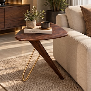 Puede incluir: Una mesa auxiliar triangular con tablero de madera oscura, una pata de madera inclinada y una pata de metal dorado en forma de horquilla. Sobre la mesa hay un libro blanco titulado The Kinfolk Home, una planta pequeña y un cuenco oscuro.