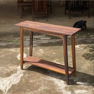 Puede incluir: Una mesa consola rectangular de madera con un patrón de vetas de color marrón oscuro y rayas naturales. La mesa tiene cuatro patas rectas y un estante inferior colocado entre las patas, ofreciendo un diseño abierto con una superficie lisa.