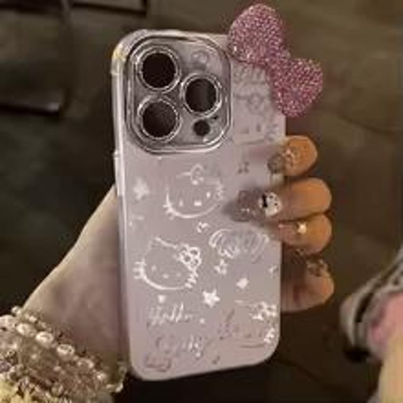 Puede incluir: Una funda transparente para tel&eacute;fono con un tinte morado claro, con dise&ntilde;os en relieve de Hello Kitty y un lazo rosa brillante. La funda tiene un protector de lente de c&aacute;mara plateado y est&aacute; sujeta por una mano con una pulsera de cuentas.