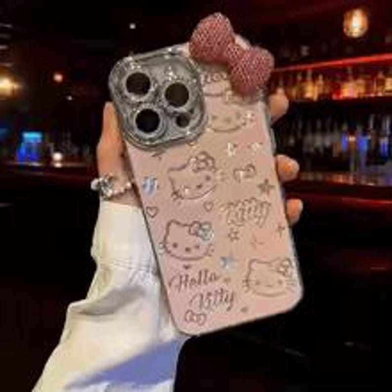 Puede incluir: Funda de tel&eacute;fono rosa con dise&ntilde;os de Hello Kitty y un lazo brillante. La funda tiene una secci&oacute;n transparente para la c&aacute;mara y est&aacute; decorada con corazones, estrellas y el texto "Hello Kitty". La funda es sostenida por una persona.