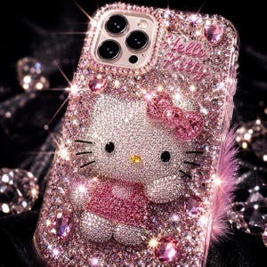 Puede incluir: Una funda de tel&eacute;fono rosa y enjoyada con un dise&ntilde;o brillante de Hello Kitty. La funda est&aacute; cubierta de peque&ntilde;os cristales brillantes e incluye un lazo rosa y las palabras "Hello Kitty". La funda tiene un borde transparente y un acento de plumas rosas.