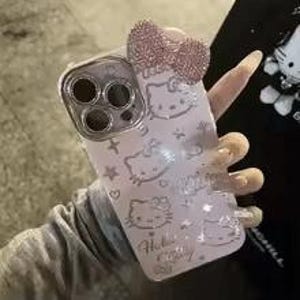 Puede incluir: Una funda de tel&eacute;fono rosa claro con dise&ntilde;os de Hello Kitty y un lazo brillante. La funda tiene un anillo de c&aacute;mara plateado y un patr&oacute;n de caras de Hello Kitty, estrellas y corazones. Las palabras "Hello Kitty" est&aacute;n impresas en la funda.