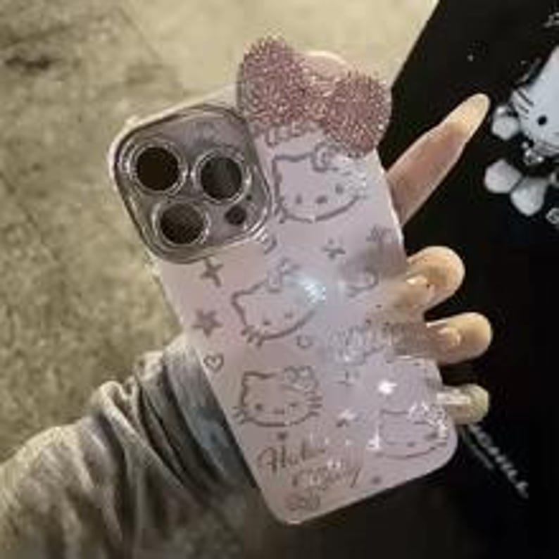 Puede incluir: Una funda de tel&eacute;fono rosa claro con dise&ntilde;os de Hello Kitty y un lazo brillante. La funda tiene un anillo de c&aacute;mara plateado y un patr&oacute;n de caras de Hello Kitty, estrellas y corazones. Las palabras "Hello Kitty" est&aacute;n impresas en la funda.