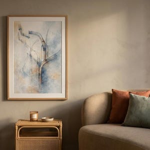 Może przedstawiać: Oprawiony abstrakcyjny wydruk artystyczny z niebieskimi i beżowymi tonami wisi nad małym stolikiem z rattanu. W pobliżu znajduje się sofa z pomarańczowymi, zielonymi i beżowymi poduszkami. Roślina doniczkowa w plecionym koszu.