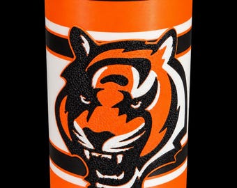 May include: A cylindrical beverage insulator featuring a vibrant orange base with horizontal white and black stripes. Centered on the front is a detailed, raised graphic of a tiger head in orange, black, and white, with a black rim at the top.