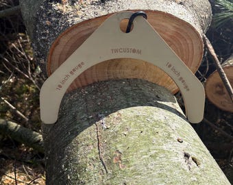 May include: A beige, triangular-shaped 10 inch gauge tool with the text TWCUSTOM printed in the center and 10 inch gauge printed on both sides, positioned over a freshly cut, light brown log with visible tree rings and bark.