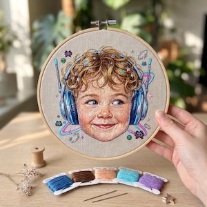 May include: A circular wooden embroidery hoop containing a detailed thread portrait of a curly-haired child wearing blue headphones. The design features small embroidered stars, pixelated space invaders, and neon-colored rings on a beige fabric.