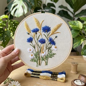 May include: A circular wooden embroidery hoop containing a fabric panel with a hand-stitched design of blue cornflowers, white daisies, and golden wheat stalks. Below the hoop are three skeins of embroidery thread in blue, white, and gold.