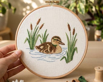 May include: A circular wooden embroidery hoop containing white fabric with a hand-stitched design of a brown duckling swimming in blue water, surrounded by green reeds and brown cattails. The embroidery features detailed thread work.