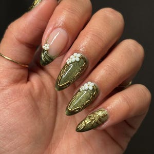 May include: Olive green nail art with gold filigree and white flower accents. The nails are almond-shaped and feature a French tip design on one nail. The design includes a gold outline and small white flowers.