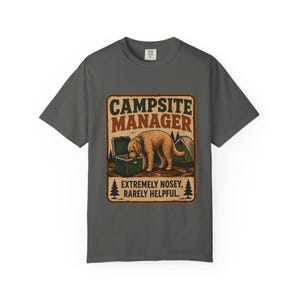 May include: Dark grey t-shirt with a graphic that reads "Campsite Manager." The graphic features a dog sniffing a cooler, a tent, and the text "Extremely Nosey, Rarely Helpful."