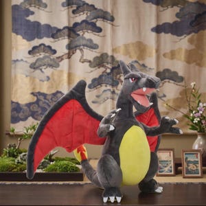 Może przedstawiać: Pluszowa zabawka Charizard z szarym korpusem, żółtym brzuchem i czerwonymi skrzydłami, w dynamicznej pozie. Smok ma ognisty ogon i otwartą paszczę. Tło z grafiką w stylu japońskim.