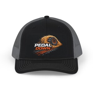 May include: Black and grey trucker cap with a curved peak. The front panel displays the words "PEDAL DOWN" in white and orange, alongside an image of a tyre track.