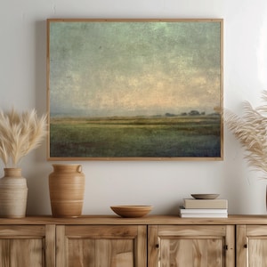May include: A rectangular landscape painting in a wooden frame featuring a muted green field under a soft, hazy sky with a textured finish. The artwork is displayed above a wooden sideboard with a ceramic vase, a bowl, and a stack of books.