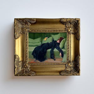 May include: A framed painting with a gold ornate frame. The painting depicts a person in a black dress reclining on a green sofa. The person is holding a yellow object. The background features green pillows.
