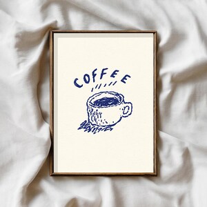 May include: A framed art print featuring a hand-drawn illustration of a coffee cup with steam rising. The word "COFFEE" is written above the cup in blue ink. The artwork is set against a cream-colored background and framed in a wooden frame.