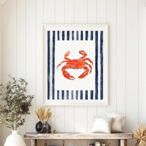 May include: Framed art print with a watercolour illustration of a red crab. The crab is on a white background, framed by navy blue vertical stripes. The artwork is in a white frame, suitable for coastal or nautical decor, measuring approximately 50 x 70 cm.