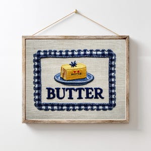 May include: Embroidered wall art featuring a butter-themed design. The artwork showcases a yellow butter pat on a blue plate, framed by a navy blue gingham border. The word "BUTTER" is embroidered below the image.