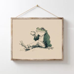 Puede incluir: Una impresión artística enmarcada que presenta una ilustración caprichosa de una rana con chaqueta y pantalones verdes, sosteniendo una taza de té. La rana está sentada, con una pierna extendida. La obra de arte es en tonos verdes, blancos y marrones.
