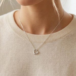 May include: A delicate silver-toned necklace featuring a small, open heart-shaped pendant suspended from a thin, polished chain. The metal has a smooth, reflective finish, and the heart design is simple, clean, and minimalist in its overall appearance.