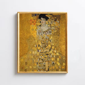 May include: A framed print of a Gustav Klimt painting. The artwork features a woman in a golden gown with intricate patterns and a black hat. The frame is gold, and the background is a rich, golden hue, creating a luxurious aesthetic.