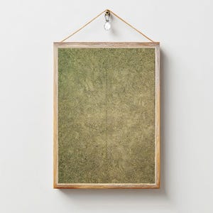 May include: A framed abstract art print with a light wood frame. The artwork features a textured pattern in shades of green and gold. The print is suspended by a natural rope and a decorative crystal. The artwork is a decorative wall hanging.