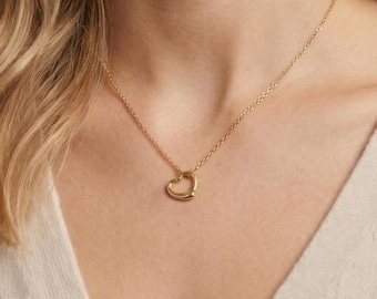 May include: A gold-toned necklace featuring a delicate chain and a small, open heart-shaped pendant. The pendant has a smooth, polished finish and is centered on the collarbone, suspended from a thin, matching gold-toned metal link chain.