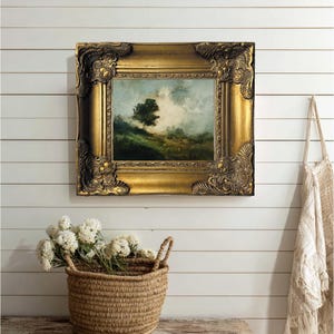 May include: A framed landscape painting with a gold ornate frame. The artwork depicts a tree on a hillside under a cloudy sky. A woven basket with white flowers sits below the painting.