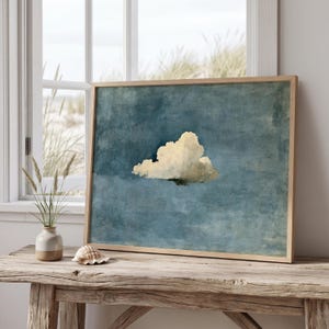 May include: A rectangular framed wall art print featuring a single, textured, off-white cloud centered against a deep, moody blue sky with visible brushstroke details. The artwork is enclosed in a light-toned wooden frame with a clean edge.