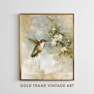 May include: A rectangular wall art piece in a thin gold frame featuring an oil painting of a hummingbird hovering near a branch of white blossoms. The background consists of soft, blended brushstrokes in shades of beige, cream, and pale blue.