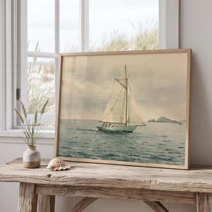 May include: A framed watercolor print depicting a sailboat with white sails navigating calm, muted blue-grey waters under a soft, overcast sky. The artwork features a distant rocky coastline and is presented in a light-toned wooden frame.