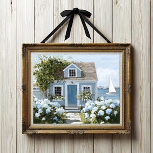 Op de afbeelding: Ingelijst kunstwerk met een kusthuisje. De schildering toont een blauw huis met een bruin dak, omringd door blauwe bloemen en groen. Een zeilboot is zichtbaar in de verte. Het kunstwerk is ingelijst in een gouden, sierlijke lijst en opgehangen met een zwart lint.
