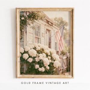 May include: A framed wall art print featuring a weathered white wooden house with a large bush of white hydrangea flowers in the foreground. An American flag hangs from the side of the building near a wooden fence, all encased in a gold frame.