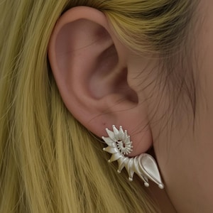 May include: A silver-toned ear cuff featuring a spiralling, shell-like design with pointed, radiating segments that curve along the outer edge of the ear. The metal has a matte, brushed finish, creating a textured appearance that follows the natural contour.