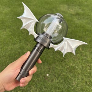 Puede incluir: Un bastón de luz gris oscuro con un globo transparente y alas de murciélago blancas. El bastón de luz tiene un mango cilíndrico y una fuente de luz cilíndrica transparente dentro del globo. Las alas están unidas al globo.