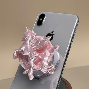 以下が含まれることがあります： 銀色のスマートフォンの背面に装着された、光沢のあるライトピンクのスマホグリップ。彫刻された翼のあるユニコーンが特徴で、詳細な翼、流れるようなたてがみ、尻尾が光沢のあるメタリック仕上げのプラスチックで表現されています。
