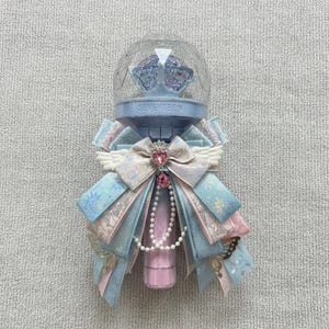 Puede incluir: Un bastón de luz decorativo azul claro y rosa con la palabra "SEVENTEEN". El bastón de luz está adornado con cintas, lazos y una cadena de perlas. La parte superior tiene un globo transparente y facetado con detalles azules.