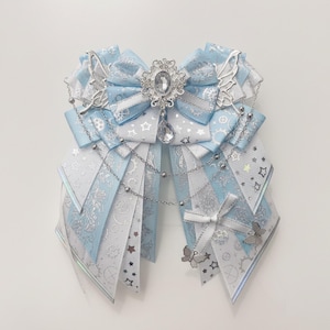 Può includere: Un fiocco per capelli a strati realizzato con nastri azzurri e bianchi con motivi celesti argentati di stelle, ingranaggi e rose. Il fiocco è decorato con una spilla ovale in strass, un cristallo pendente, ciondoli ad ala e catenelle.